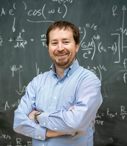 Man in a light blue dress shirt standing in front of a chalkboard with equations with his arms crossed
