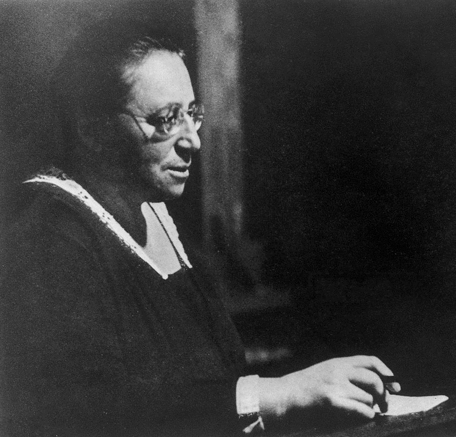 B&W photo of Noether in profile holding a pen over paper, with dark hair pulled back, wearing a simple dark dress with light embellished edges and round thin glasses.