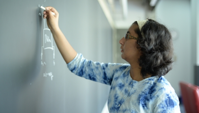 Side profile of a woman wearing a blue tie dye shirt writing on a blackboard