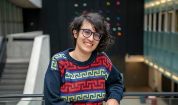 A young woman with short curly dark hair and glasses with a patterned sweater standing in an atrium